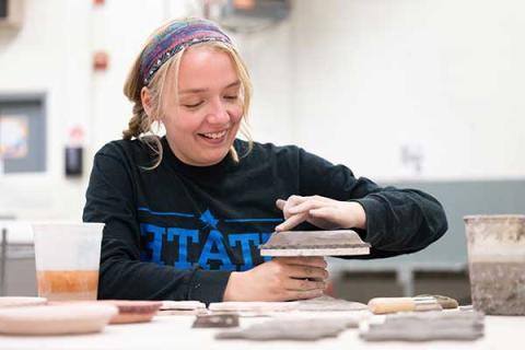 A young, smiling blonde woman, wearing a multicolored scarf in her hair and a navy blue sweatshirt that says STATE in royal blue letters, rubs her finger along the top of an unfinished clay disc on a potter’s wheel. Other flat clay items are visible on the table in front of her, and a bucket of clay can be seen to her right.