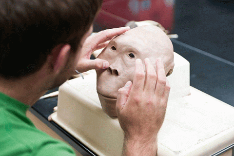 A white male with short brown is working on a 3D model reconstruction of a white human face and head. His hands are on the model, which is positioned on a white surface. 